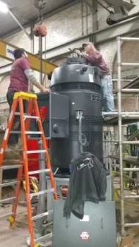 Two workers are atop ladders, servicing a large grey machine in an industrial setting, with scaffolding and overhead equipment visible