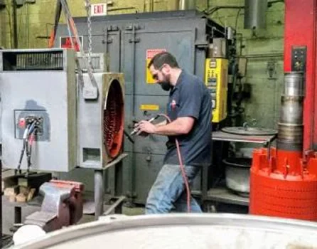 A worker uses a pneumatic tool in a workshop, surrounded by machinery and equipment, focusing on a metal piece. Safety signs are visible