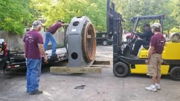 Four workers assist in unloading a large, round machine part using a forklift in a wooded outdoor setting