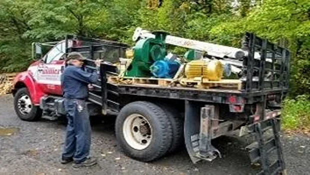 A worker inspects a flatbed truck loaded with industrial equipment, including motors and machinery, in a wooded outdoor setting