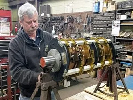 A person stands in a workshop holding a large, complex mechanical assembly with metallic components and a circular design
