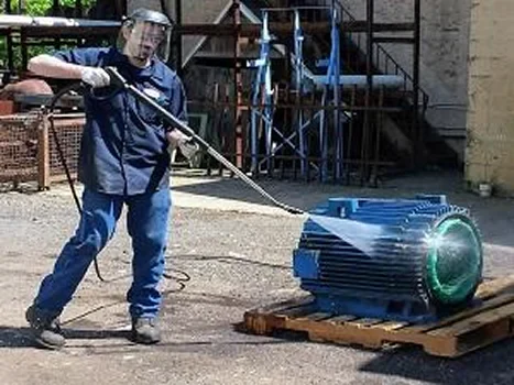 A worker uses a pressure washer to clean a large electric motor on a wooden pallet, surrounded by industrial equipment
