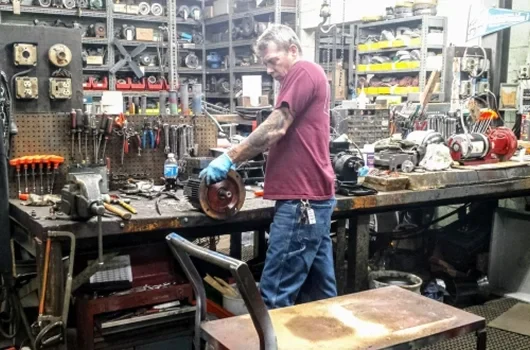 A person in a workshop inspects a mechanical part, surrounded by tools and equipment, with a cluttered workbench in the background