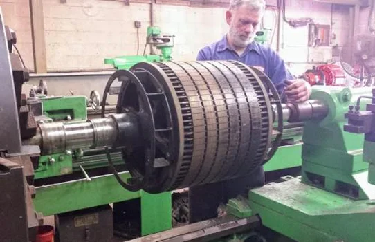 A worker operates a large industrial lathe, shaping a cylindrical metal part with multiple grooves on its surface
