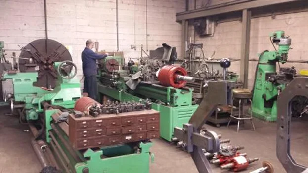 A machinist operates a lathe in a spacious workshop filled with various green machinery and tools