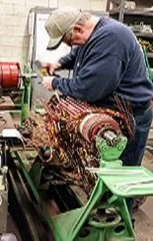 A man in a cap repairs an electric motor on a workbench, focused on rewiring its intricate copper coils and connections