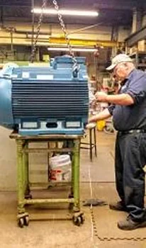 A worker inspects a large blue electric motor on a green cart in a workshop, with tools and equipment visible in the background
