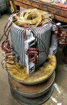 A partially disassembled electric motor with visible coils and metal components, resting on a circular metal base in a workshop setting