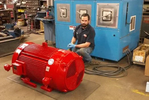 A worker in a workshop examines a large red electric motor beside a blue industrial testing machine. Tools and equipment are visible in the background