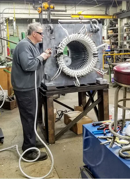 A technician works on a large industrial machine with coils and pipes, surrounded by tools and equipment in a workshop setting