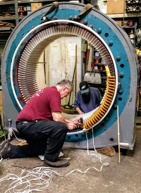 Two engineers work on a large industrial machine's coil, focusing on intricate wiring and assembly in a workshop setting
