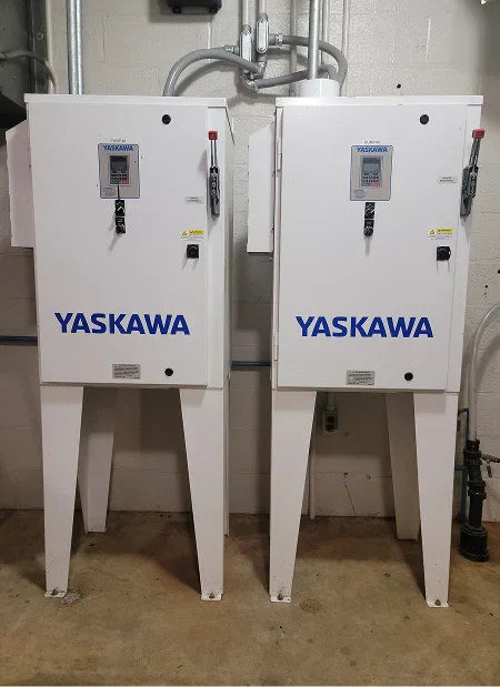 Two Yaskawa control cabinets with digital displays and red emergency stop buttons, mounted on legs in a utility space